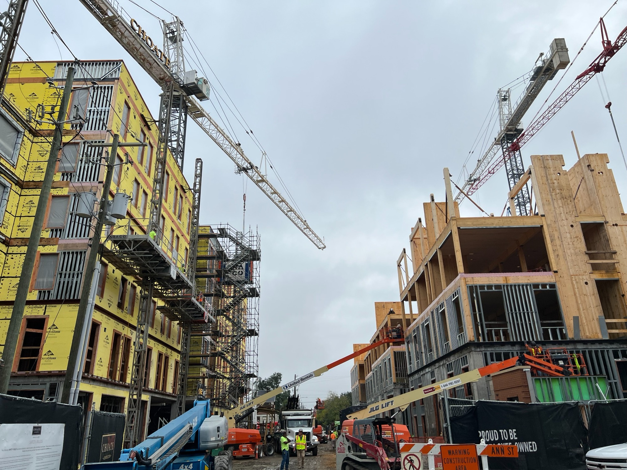 Two buildings under construction along a street, with cranes and lifts between them