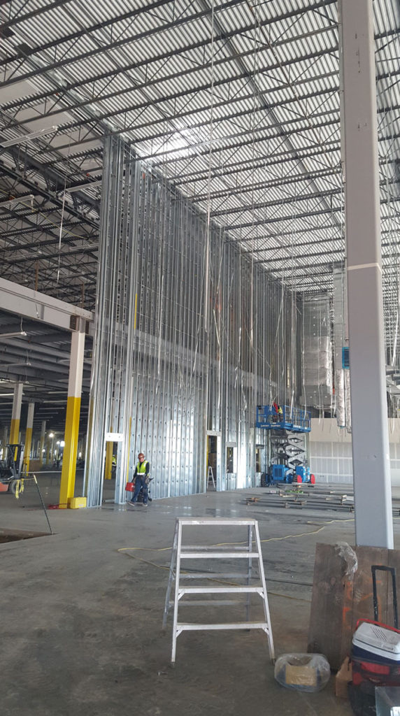 A tall metal-stud wall inside a wide industrial building, with a worker and a lift